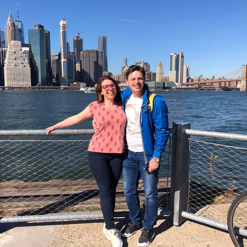 Two people are smiling for the camera with a backdrop of the New York City skyline and the Brooklyn Bridge