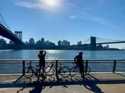 Two people with bicycles are standing by a river railing, with a city skyline and two bridges in the background, the scene illuminated by sunlight sparkling on the water.