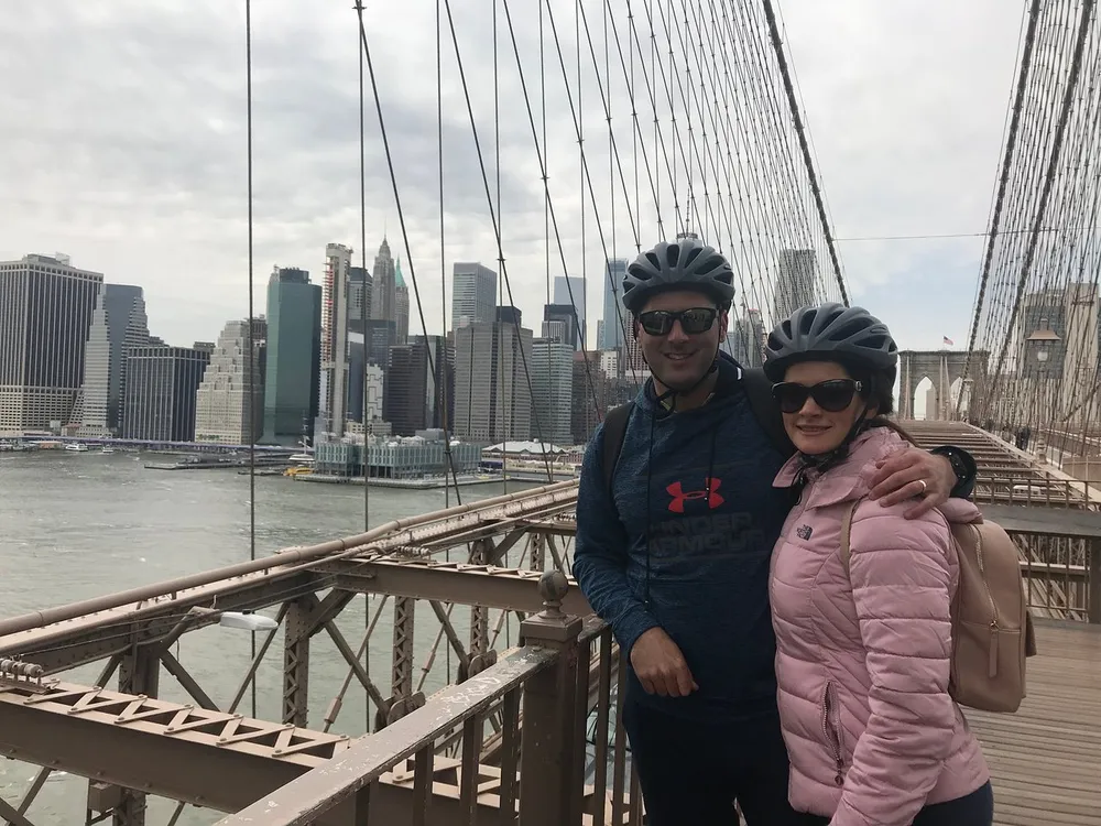 A smiling couple wearing bike helmets poses for a photo on the Brooklyn Bridge with the Manhattan skyline in the background