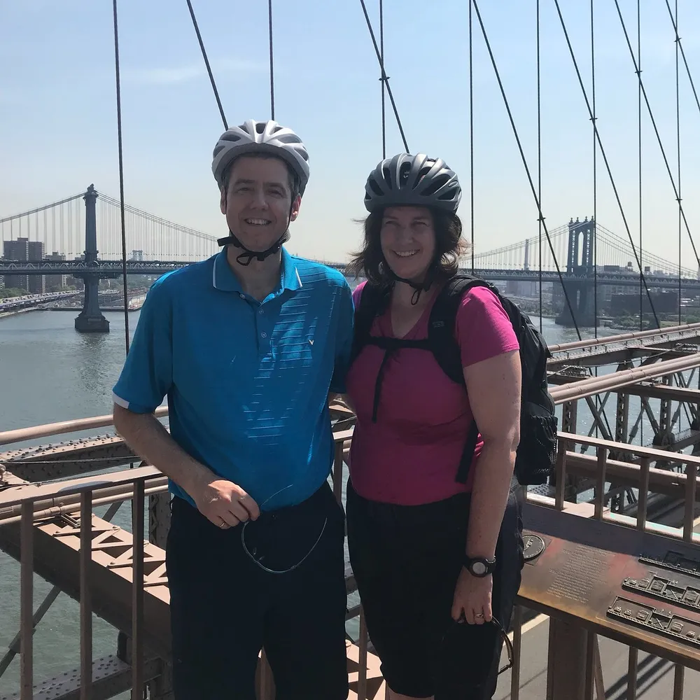 Two people wearing cycling helmets are smiling for a photo on a bridge with a scenic view of another bridge and a city skyline in the background