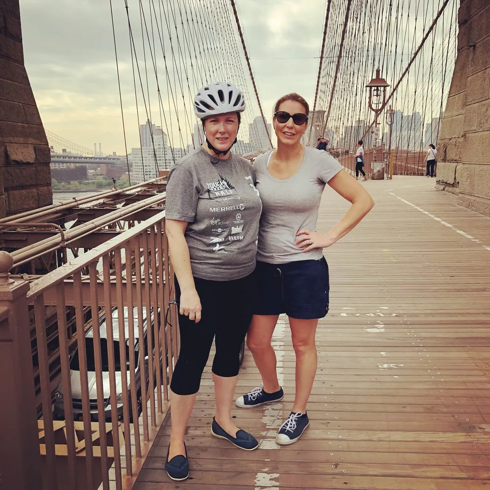 Two women are posing for a photo on what appears to be the Brooklyn Bridge with one of them wearing a bike helmet and casual activewear while the other is dressed in a casual t-shirt and shorts
