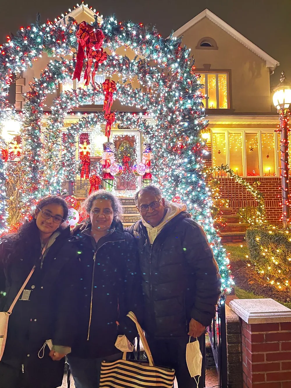 Three people are posing for a photo in front of a festively decorated house with a large illuminated Christmas wreath archway