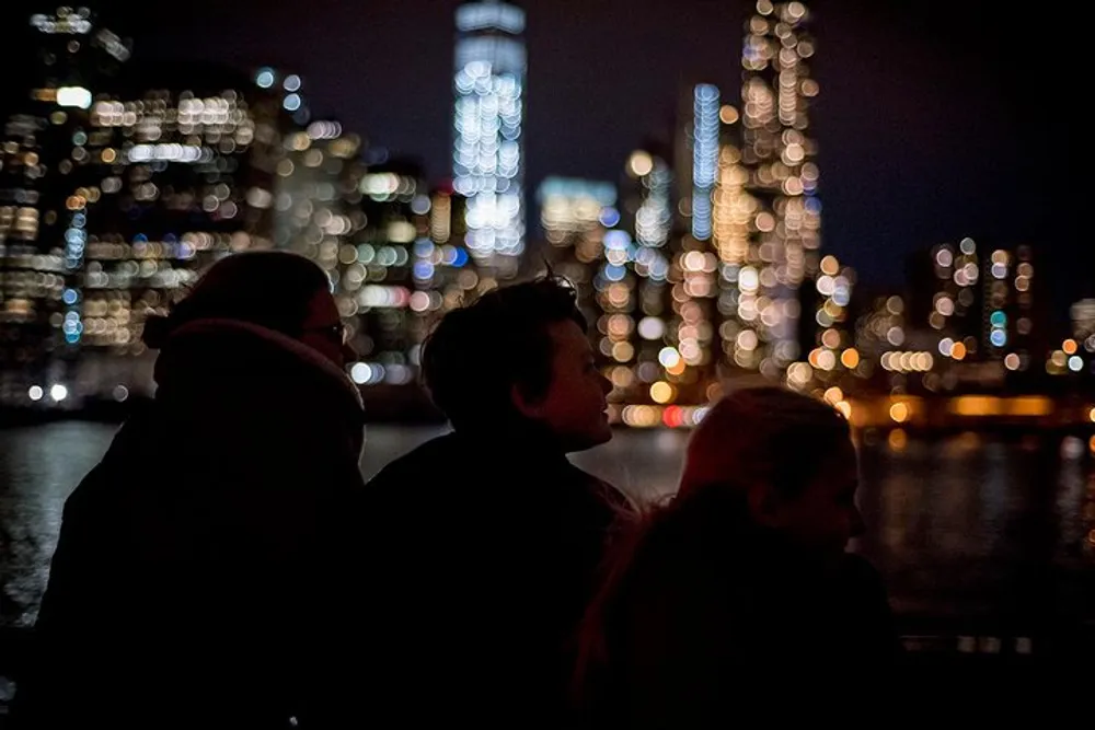Three silhouetted individuals are gazing at a brightly lit city skyline at night across a body of water