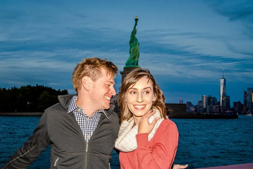 A smiling couple poses for a photo with the Statue of Liberty and the New York City skyline in the background during twilight