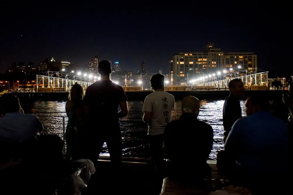A group of people stand by the water at night looking towards a cityscape illuminated by lights