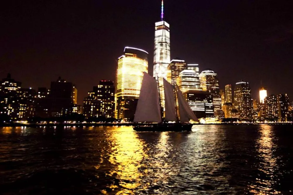 A sailboat is gliding across the water at night with the brightly lit skyline of Lower Manhattan in the background