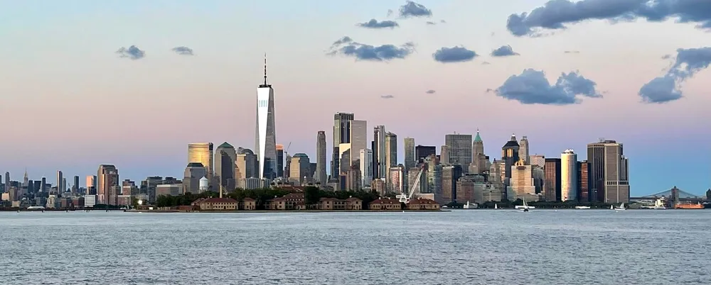 The image features a panoramic view of the Lower Manhattan skyline including the One World Trade Center as seen across the water during dusk with a soft pink sky in the background