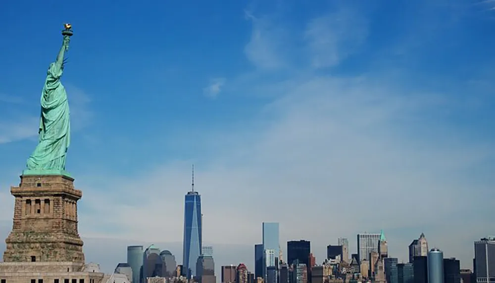 The Statue of Liberty stands prominently in the foreground with the skyline of Lower Manhattan including One World Trade Center in the background under a clear blue sky