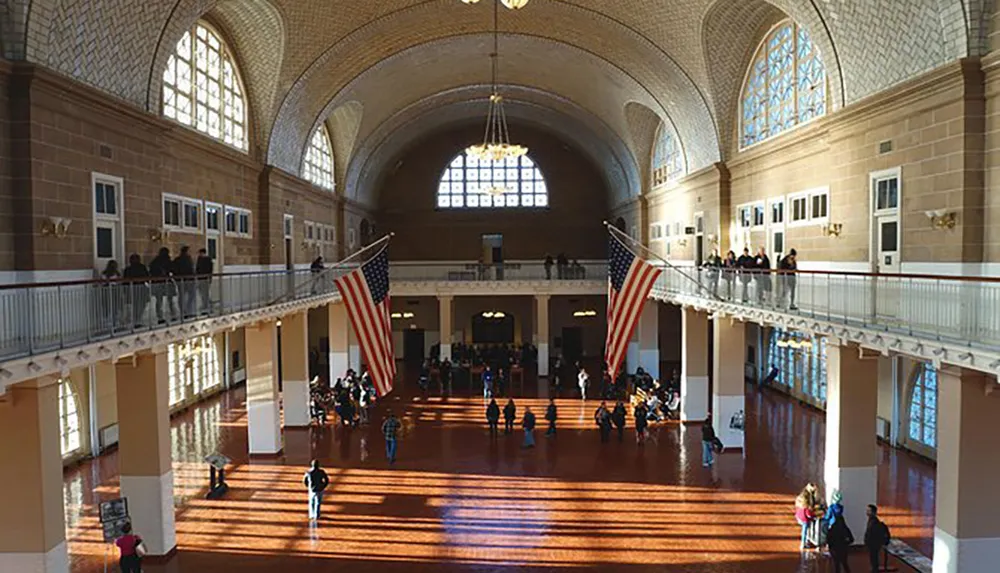 The image shows a spacious hall with a high arched ceiling large American flags hanging from the balcony and people scattered throughout the floor and balcony likely a historical building with a significance to immigration