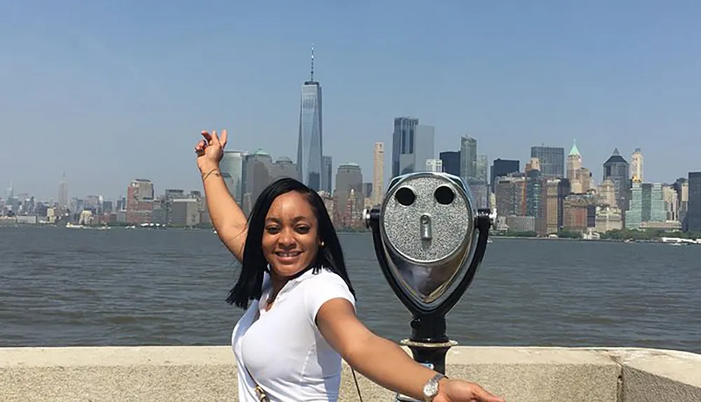 A person poses with an outstretched arm appearing to touch the top of a skyscraper in the distance with a view of a city skyline and a coin-operated binocular in the foreground