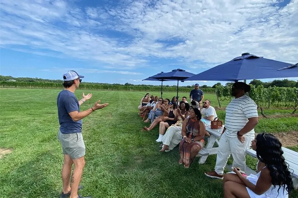 A person is giving a presentation to a group of attentive listeners seated outdoors under umbrellas with a vineyard in the background