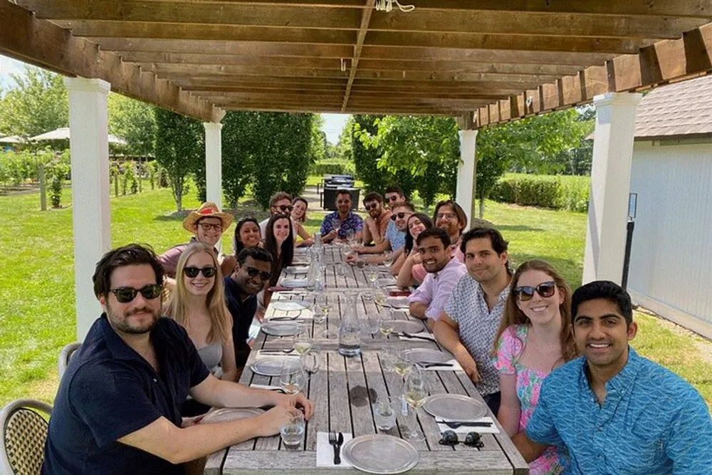 A group of smiling people are seated around a long table outdoors ready for a communal dining experience under a pergola