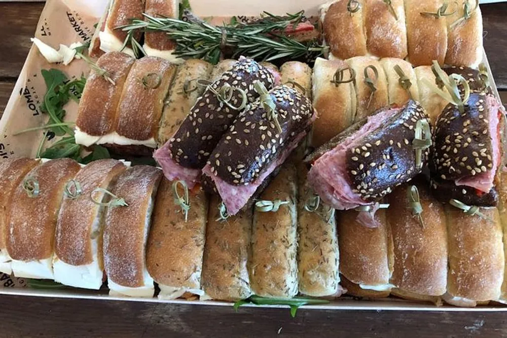 The image shows a variety of gourmet sandwiches on a tray garnished with fresh herbs
