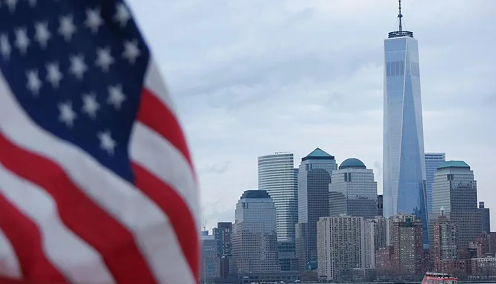 The image shows an out-of-focus American flag in the foreground with a clear view of the Lower Manhattan skyline including the One World Trade Center in the background