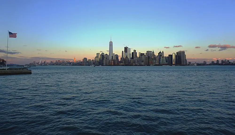 The image captures a serene sunset view of the New York City skyline with the prominent One World Trade Center standing tall viewed from across the water