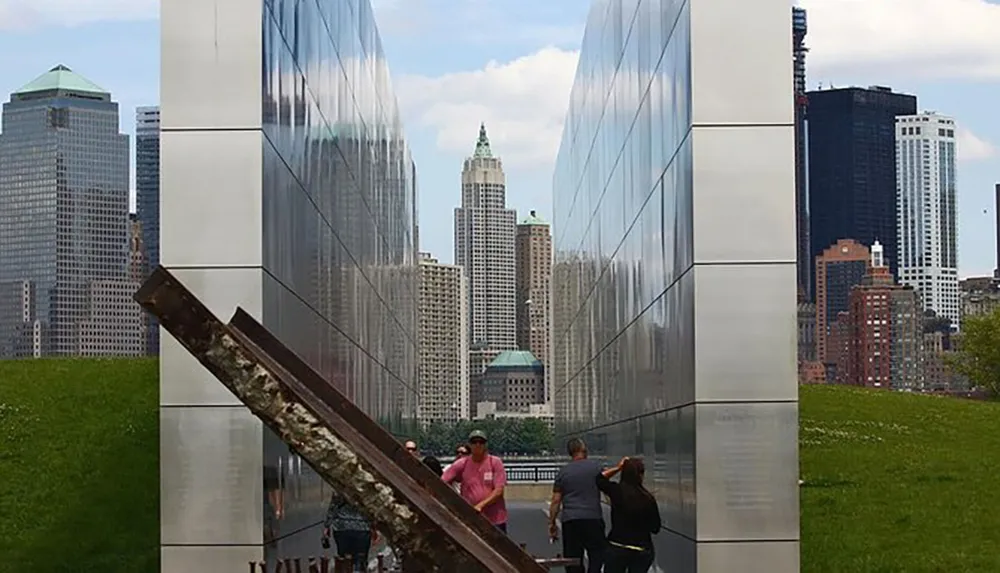 This image shows people visiting a reflective memorial installation with a piece of rusted metal in the foreground and the skyline of a city in the background
