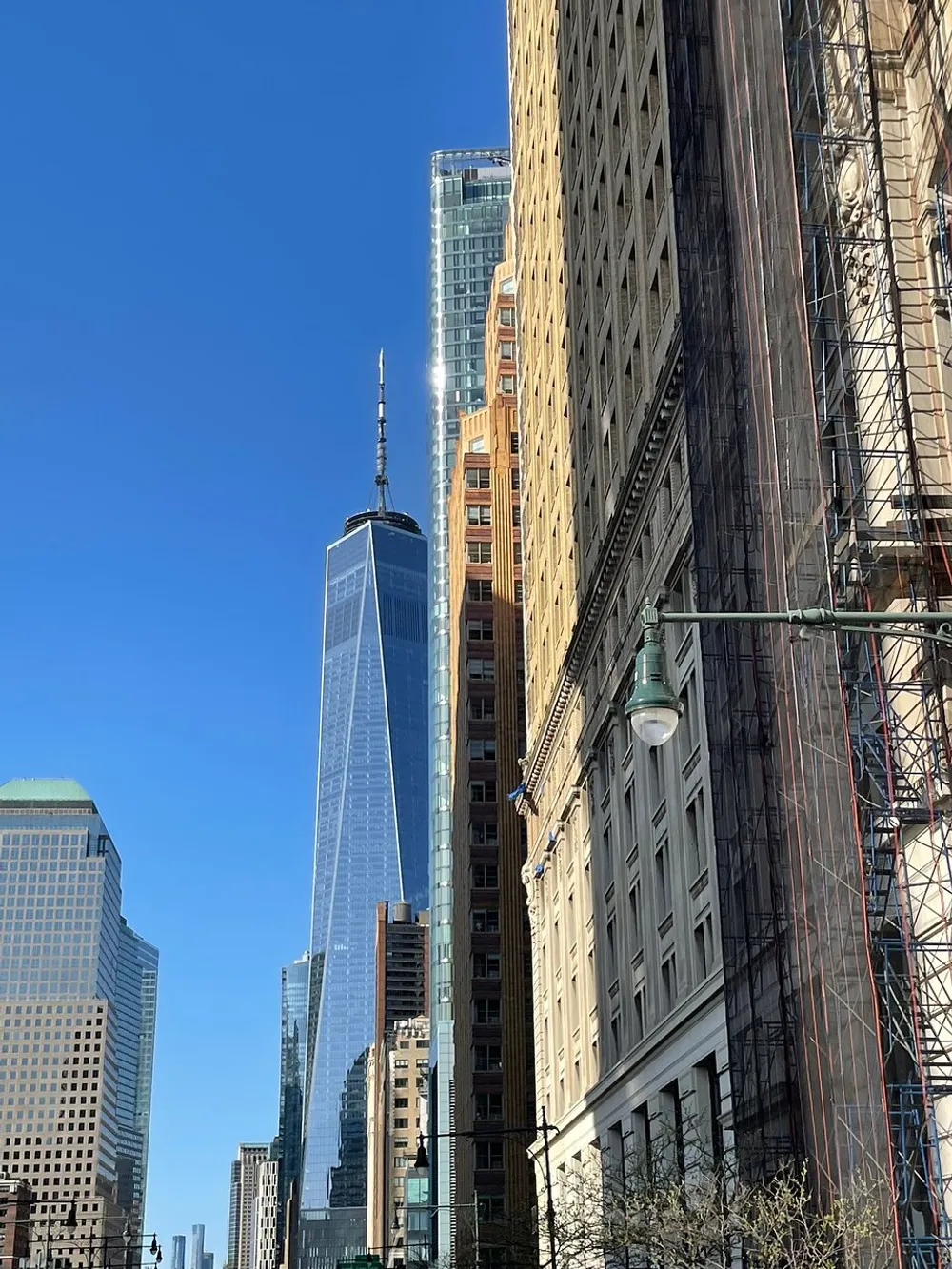 The image shows a view of the One World Trade Center amidst surrounding buildings against a clear blue sky with evidence of construction or renovation on the building in the foreground