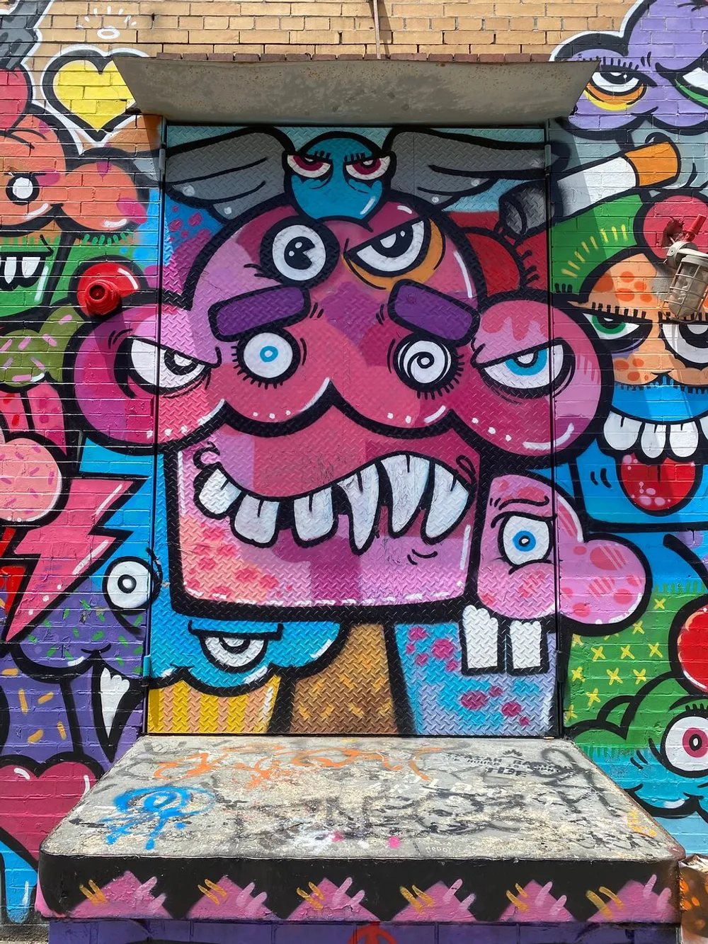 The image shows a colorful and vibrant graffiti mural of whimsical creatures with multiple eyes and expressive faces painted on an urban wall