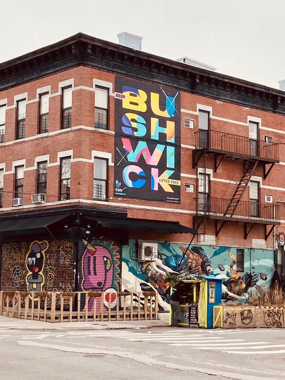The image shows an urban street corner with a red brick building adorned with vibrant street art including a large mural spelling BUSHWICK with more colorful graffiti and a food vendor stand at street level