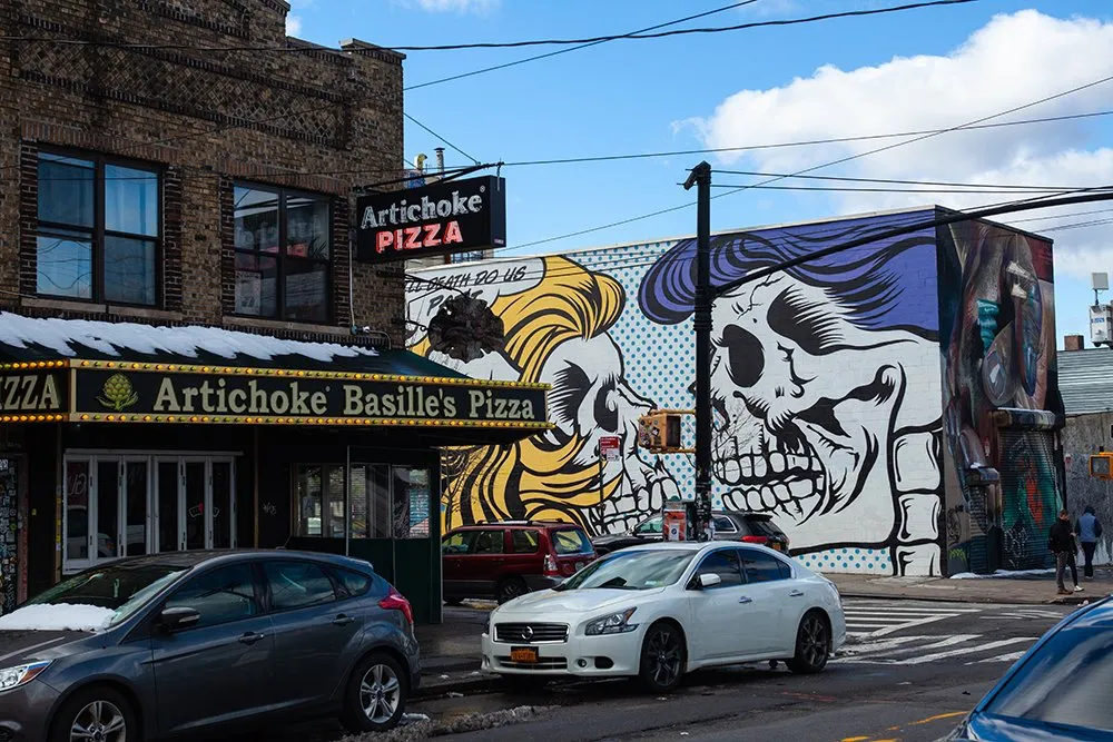 A bustling street corner featuring Artichoke Basilles Pizza restaurant with a large dramatic mural of cartoon-like figures on an adjacent buildings wall