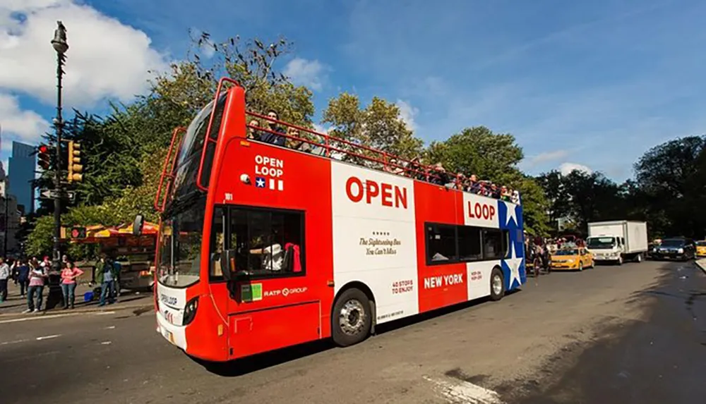 A red double-decker sightseeing bus is driving through a bustling city street likely in New York with passengers on the open top deck enjoying the view