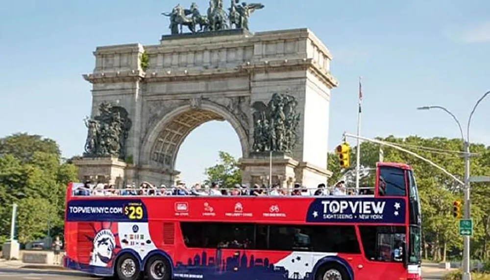 A red double-decker sightseeing bus is passing by a grand arch monument with tourists on the open-top upper deck enjoying the view