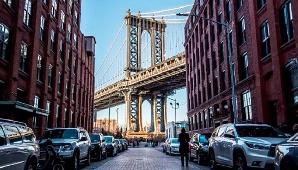 The image showcases a classic view down a cobbled street flanked by brick buildings with the iconic steel structure of a suspension bridge towering in the background aligned with the street and a person walking toward the camera