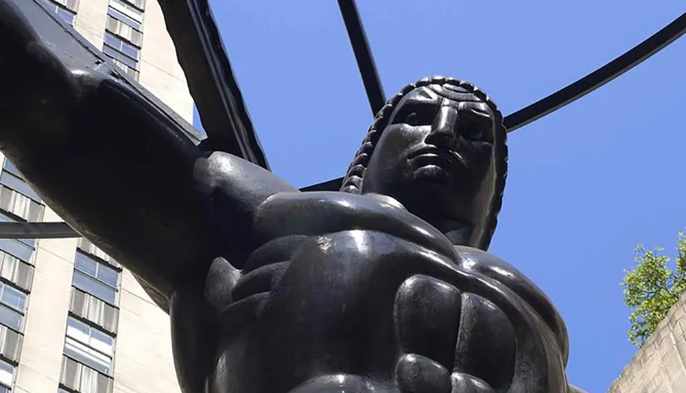 The image captures a view looking up at a large dark bronze statue of a muscular man with defined features and an outstretched arm against a clear sky