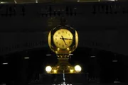 The image shows an iconic golden clock, illuminated and prominently displayed indoors, with the inscription 