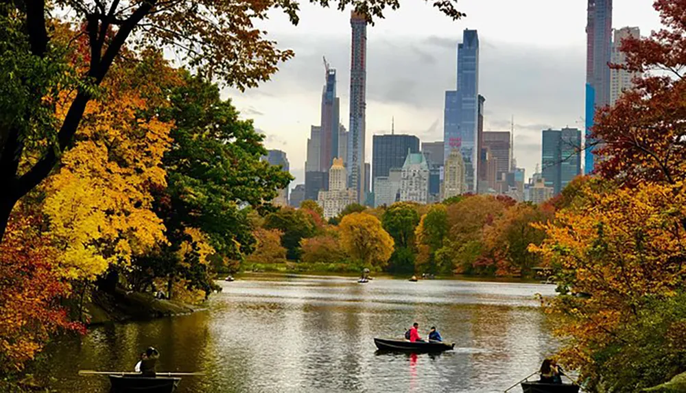 People enjoy boating on a calm lake against a backdrop of colorful autumn trees and the contrasting skyline of a bustling city