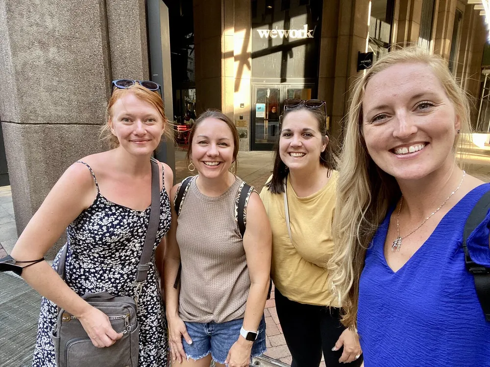 Four smiling women are posing together for a photo on a city street with one of them holding the camera for a selfie