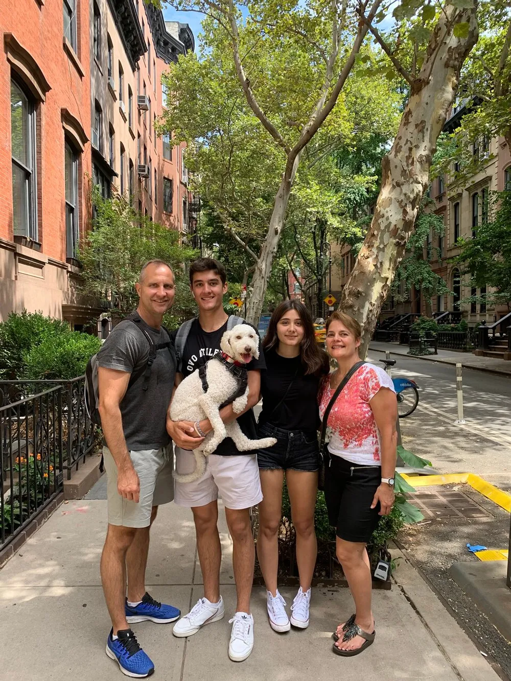 A happy family of four and their dog posing for a photo on a tree-lined urban sidewalk