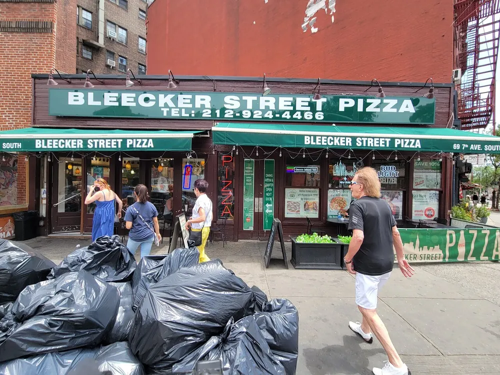 Several people are walking past a pizza shop named Bleecker Street Pizza on a sunny day with a pile of black garbage bags in the foreground
