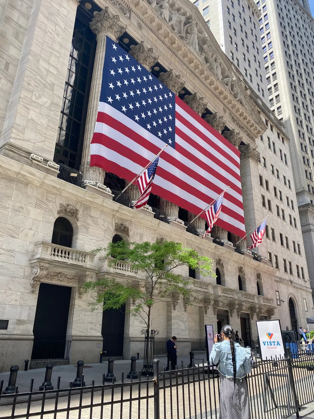 The image depicts a large American flag hanging from the facade of a stately building with Classical architecture as passersby walk in front