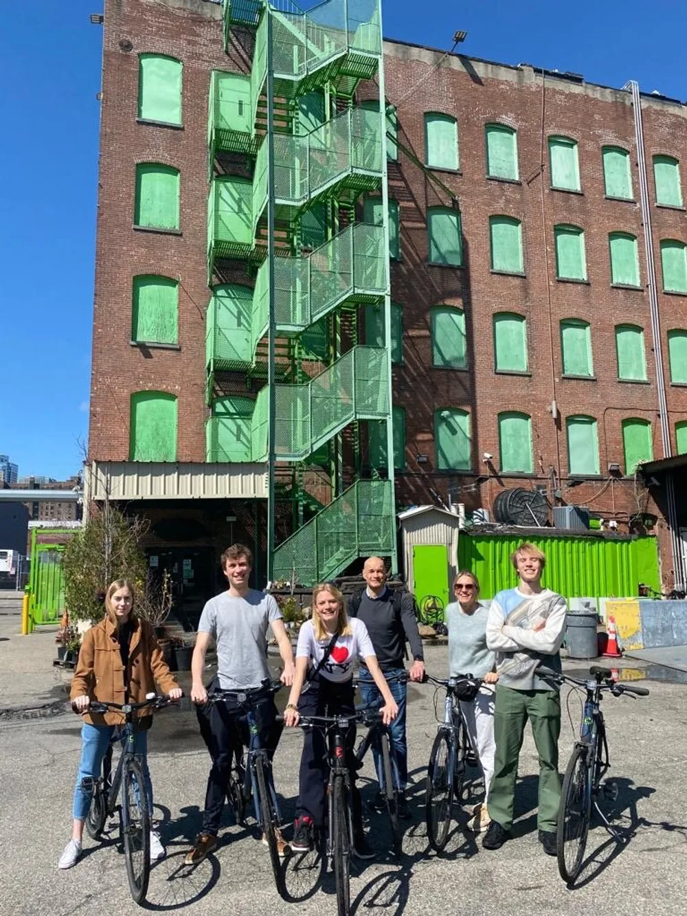 A group of six individuals are standing with bicycles in front of a brick building with a green fire escape under a clear blue sky