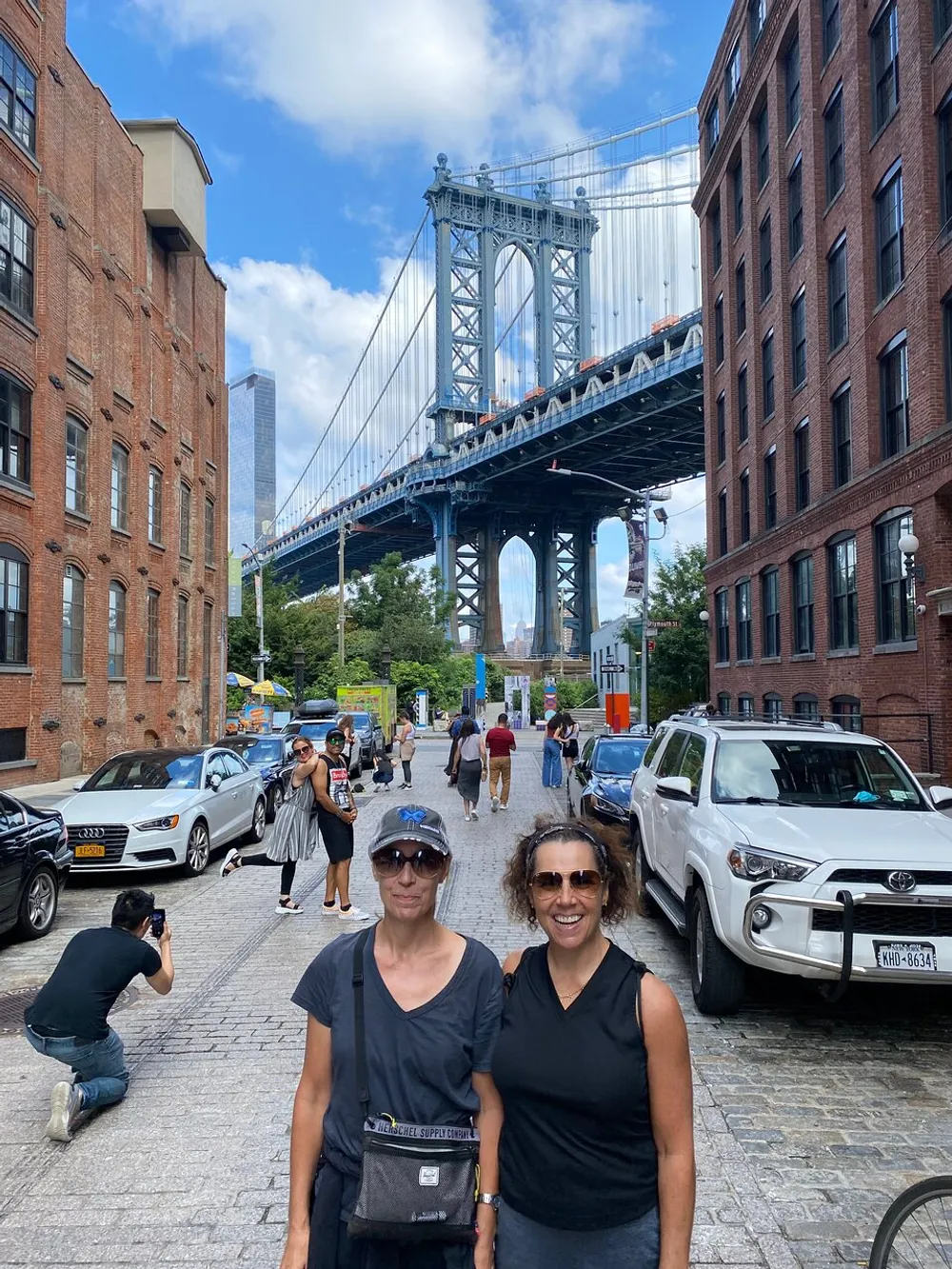 Two individuals pose for a photo on a cobblestone street with the Manhattan Bridge towering in the background amidst pedestrians and vehicles under a partly cloudy sky