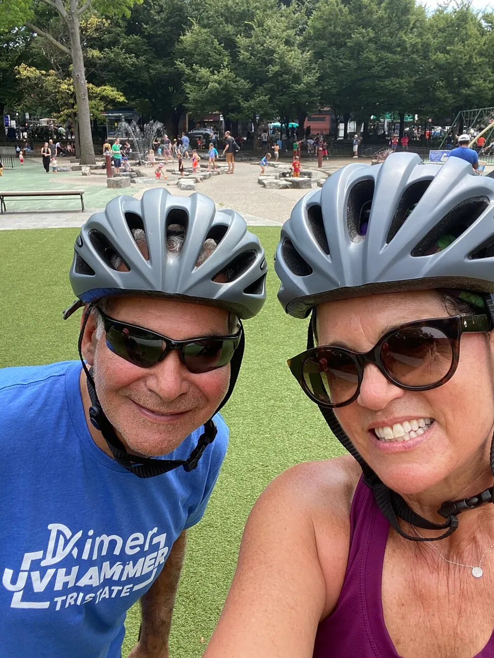 Two people wearing bicycle helmets are taking a selfie together with a background of an outdoor area where children are playing in a water feature