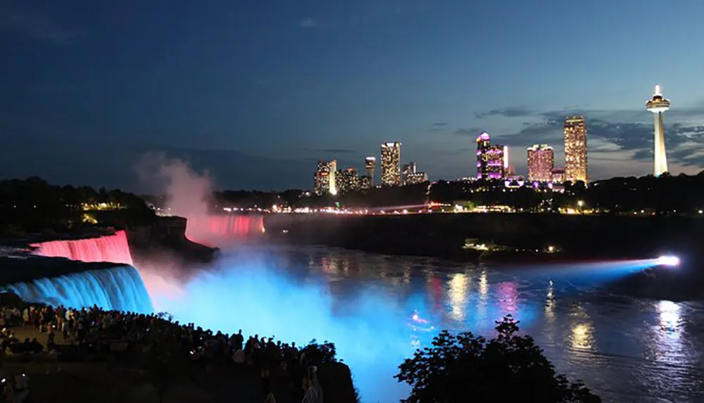The image captures a night view of Niagara Falls with illuminated water in blue and red overlooking a lit city skyline and gathering of spectators