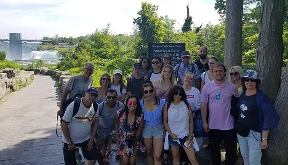 A group of people poses for a photo in front of a Niagara Falls State Park sign at a viewpoint with the falls partially visible in the background
