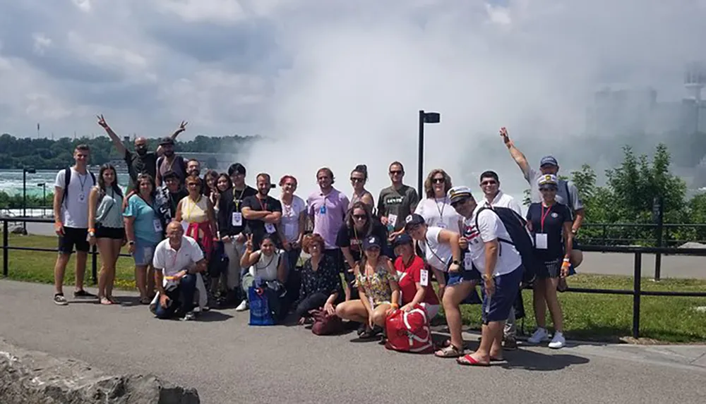 A group of cheerful people is posing for a photo with the mist from a large waterfall in the background suggesting they are visiting a famous natural attraction