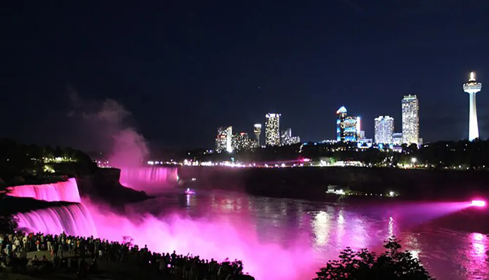 Niagara Falls is illuminated in vibrant pink lights at night with a backdrop of the city skyline and a gathering of spectators enjoying the view