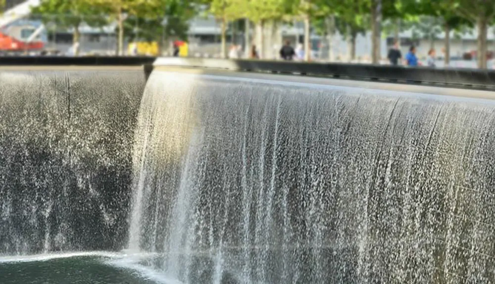 A cascading urban waterfall is in the foreground with blurred views of pedestrians and a tree-lined street in the background