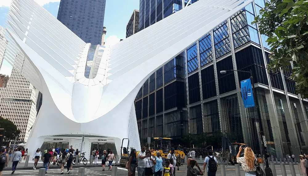 The image shows a bustling urban scene with people walking in front of the distinctive modern white ribbed structure of the Oculus in New York City