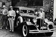 Three men in vintage clothing, two wielding firearms, stand beside a classic car with a 1930s-style design in front of a brick building.