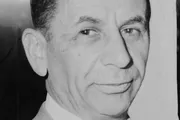 The image shows a close-up of a smiling gentleman with a part of his face obscured, presented in black and white.