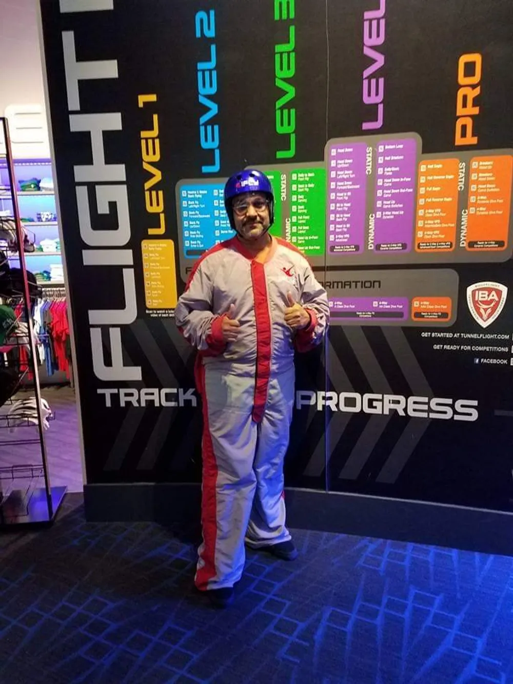A person is giving a thumbs-up while wearing a flight suit and helmet standing in front of a wall that shows various levels of flight skills