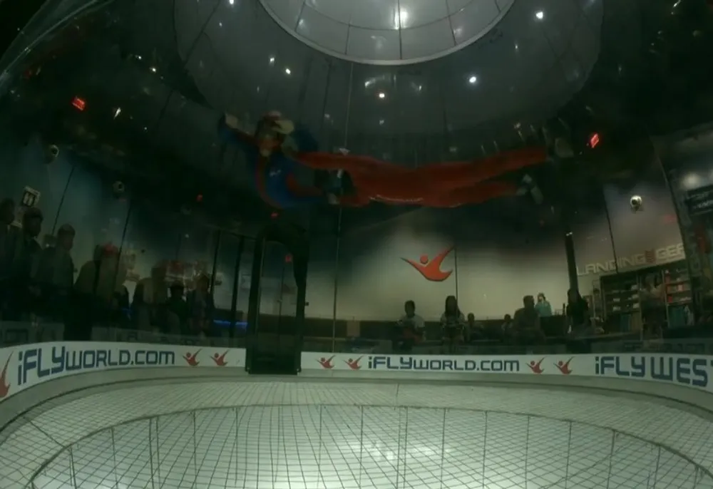 A person is indoor skydiving in a vertical wind tunnel floating in the air with a long red fabric trailing behind