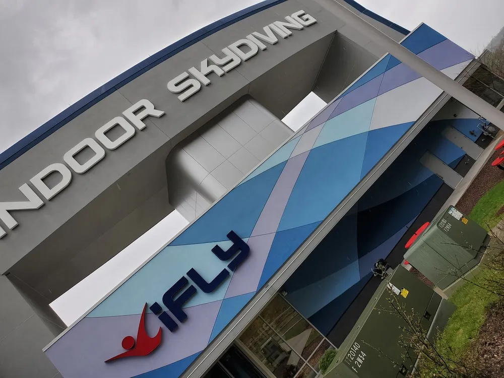 The image shows the exterior of an iFLY Indoor Skydiving facility with a modern design and the brand name prominently displayed on the building
