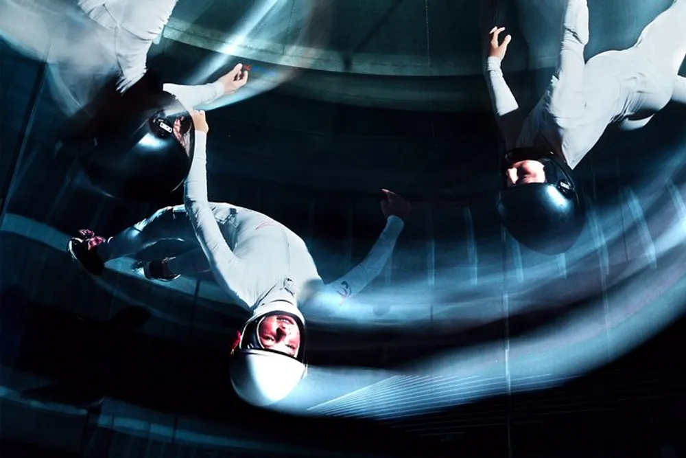 The image depicts individuals in white suits and helmets seemingly floating or flying within a wind tunnel captured with a motion blur effect
