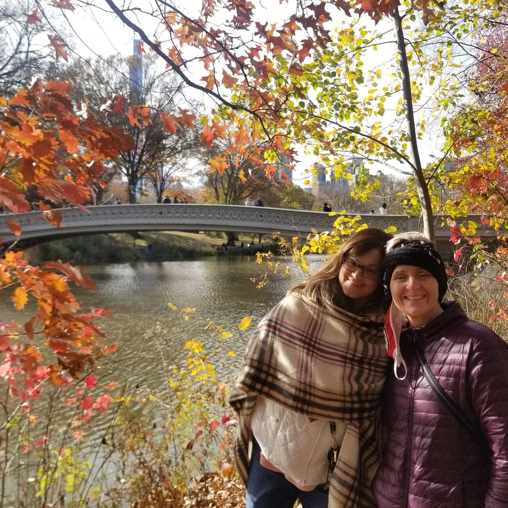 Two smiling individuals posing for a photo in a park with autumn foliage and a bridge in the background